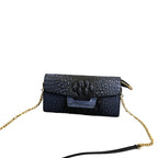 Black crocodile-patterned handbag with gold chain on a gray surface with books in the background